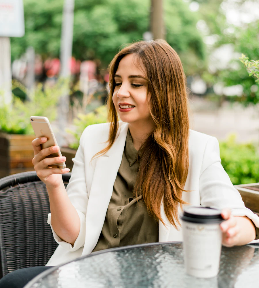 Uma mulher de blazer claro sorri ao ler uma proposta no celular, sentada à mesa de um café ao ar livre.