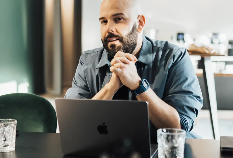 A bearded man, hands clasped in front of his laptop, listens closely during a work conversation.