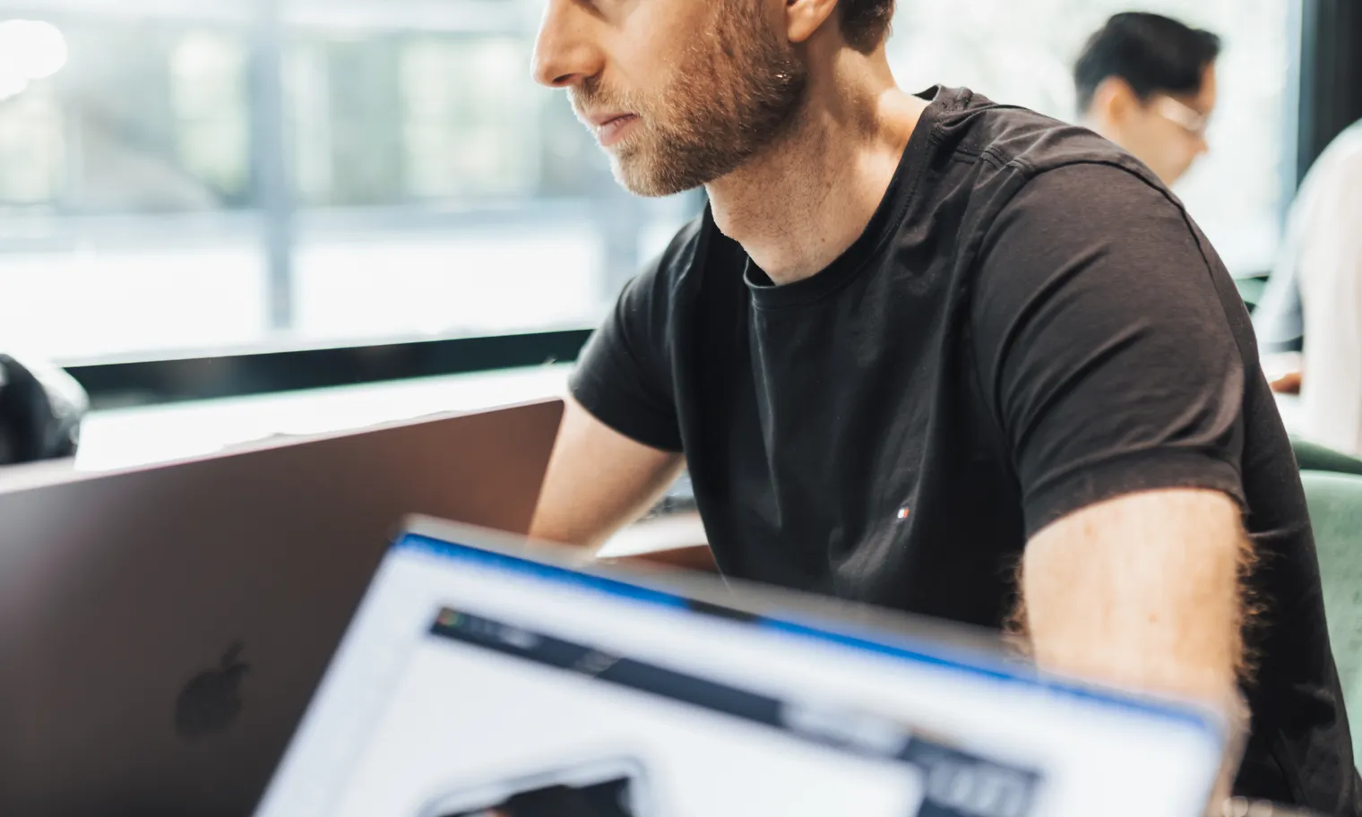 A colleague, focused, works on a laptop at a shared table.