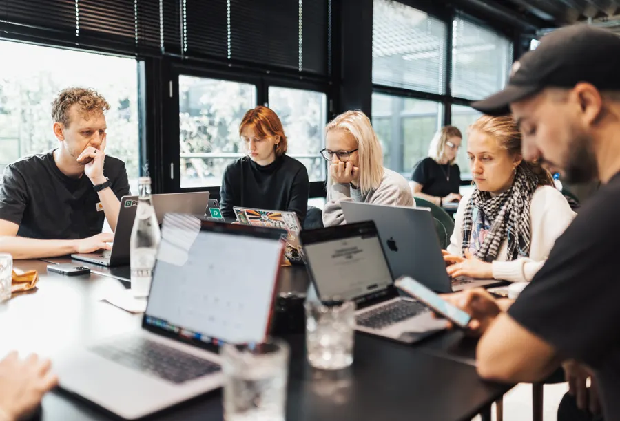 People from different teams work side by side on laptops at a long table, in front of large windows.