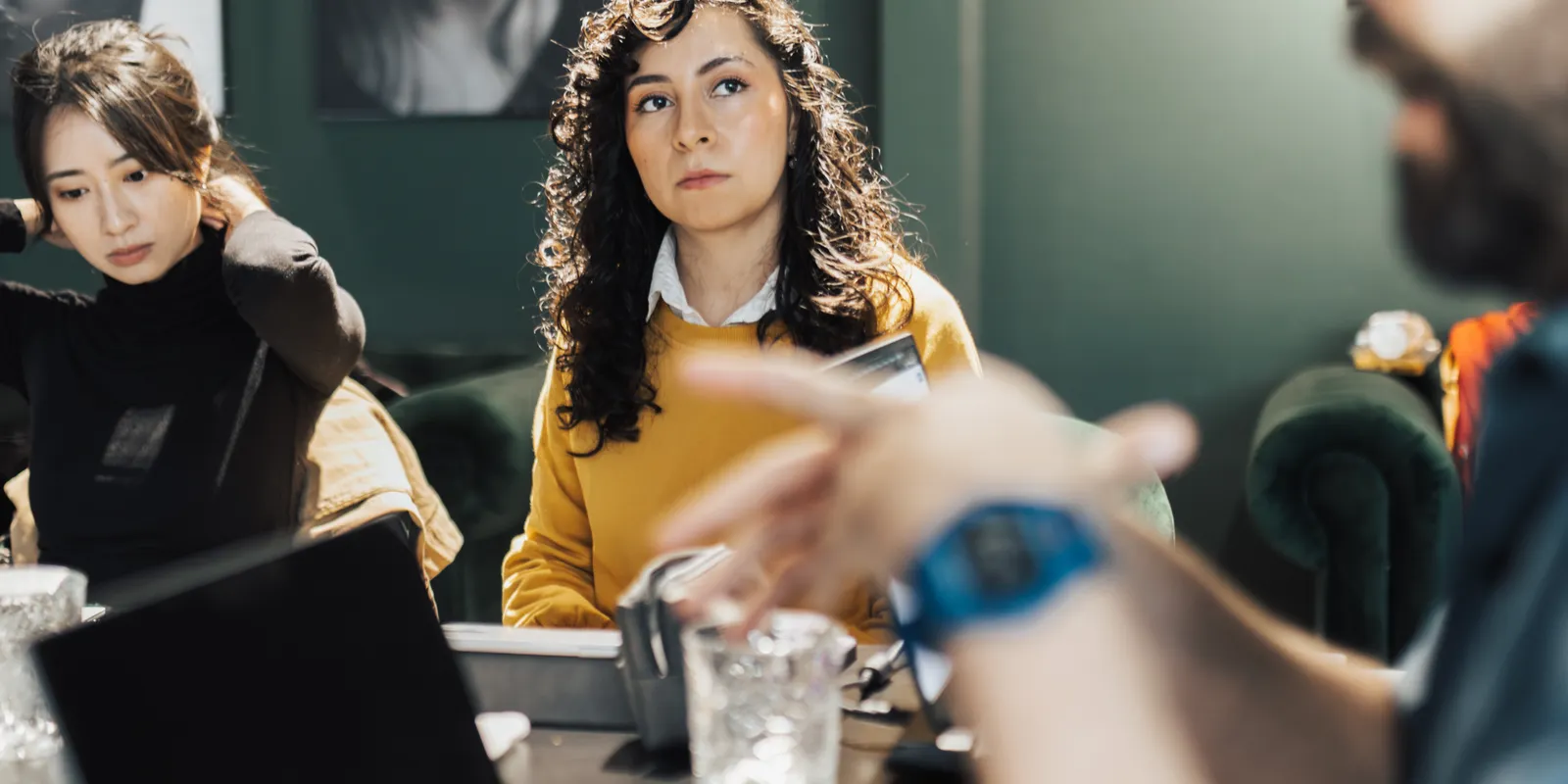 Three people sitting at a meeting room table listen to someone gesturing as they explain something.