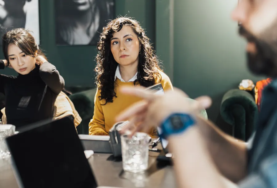 Three people sitting at a meeting room table listen to someone gesturing as they explain something.