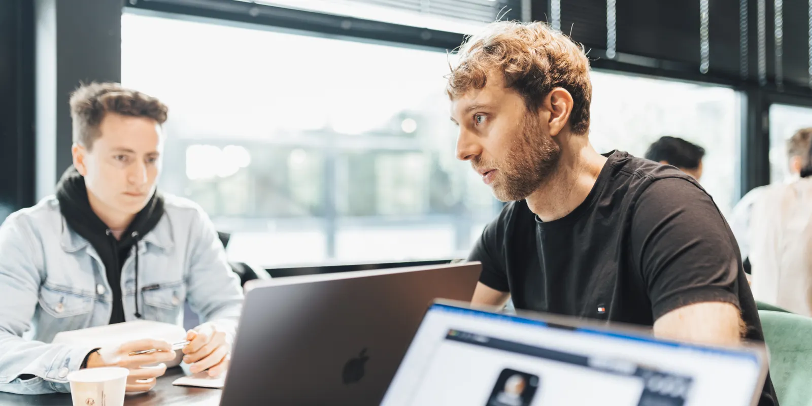 Two colleagues talk in front of an open laptop at a table beside large windows.