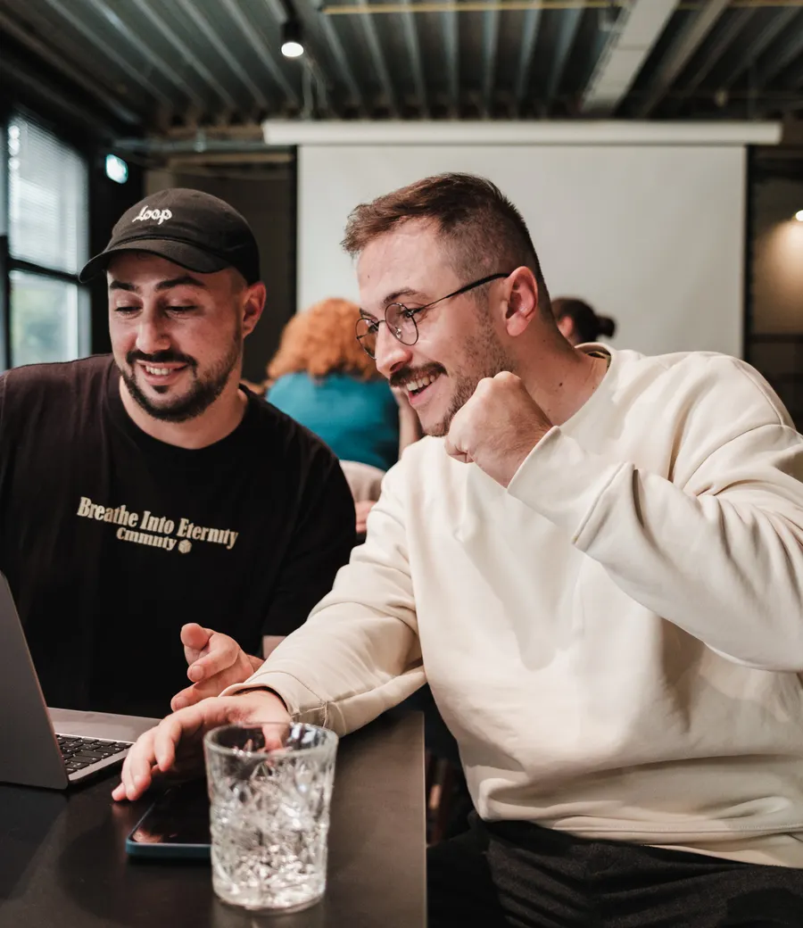 Dois programadores, um de boné e outro de óculos, riem enquanto trabalham lado a lado com os notebooks.