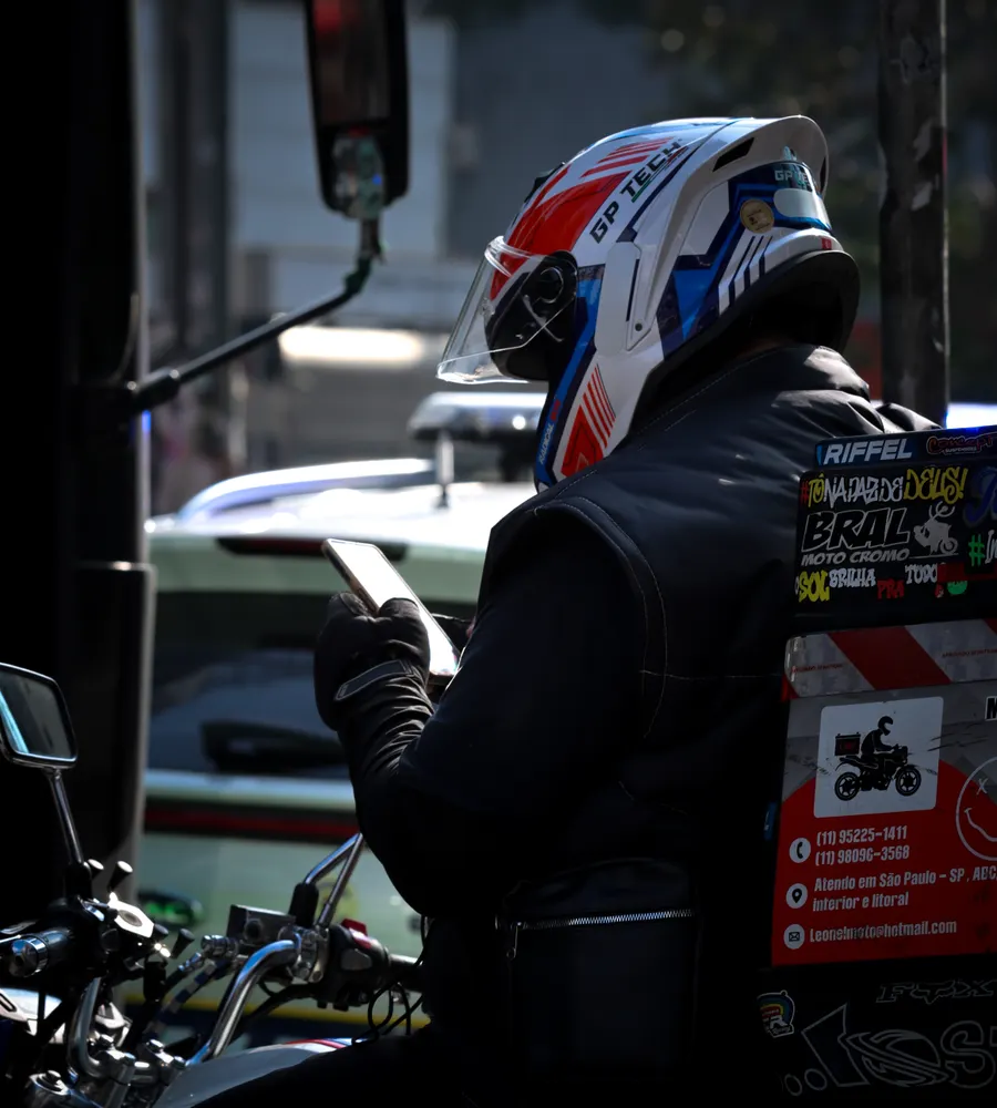Motociclista de capacete e colete, parado no trânsito de São Paulo, olhando o celular.