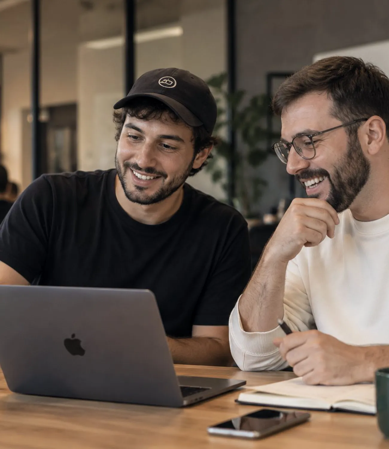 Dos programadores, uno con gorra y otro con gafas, se ríen mientras trabajan codo con codo con sus portátiles.